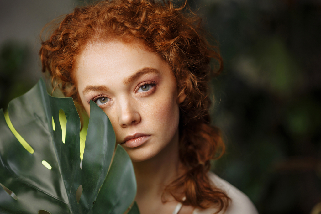 Close-up face of a beautiful young woman with red hair and freckles covering her face with a green monstera leaf while looking at the camera. Portrait of a woman with natural make-up.